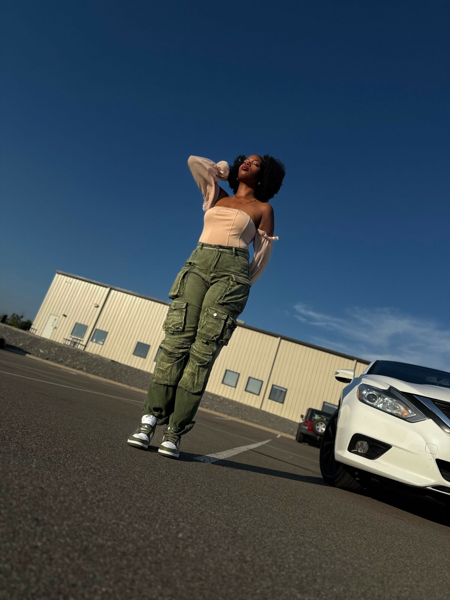 Person standing outdoors in a parking lot, with a building and a white car nearby, under a clear blue sky.