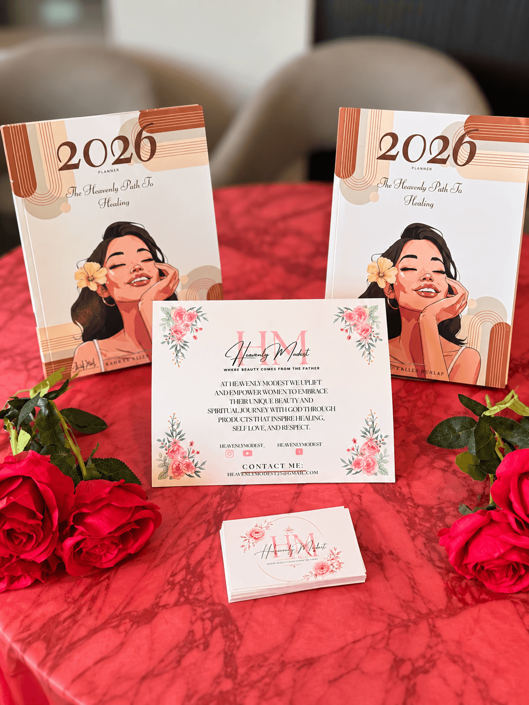 Two 2026 calendars, a floral-themed invitation, and a small card on a red table with roses, in a room setting.