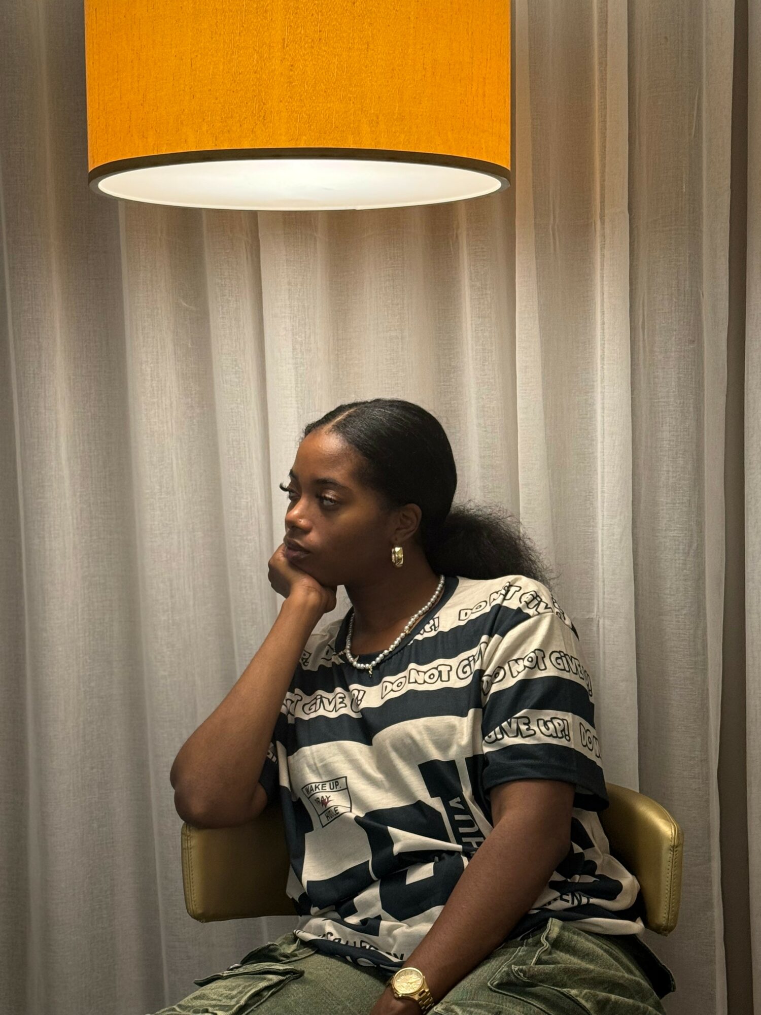 Woman sitting in a chair under a hanging orange lampshade, with curtains behind her, wearing a patterned shirt and jewelry.
