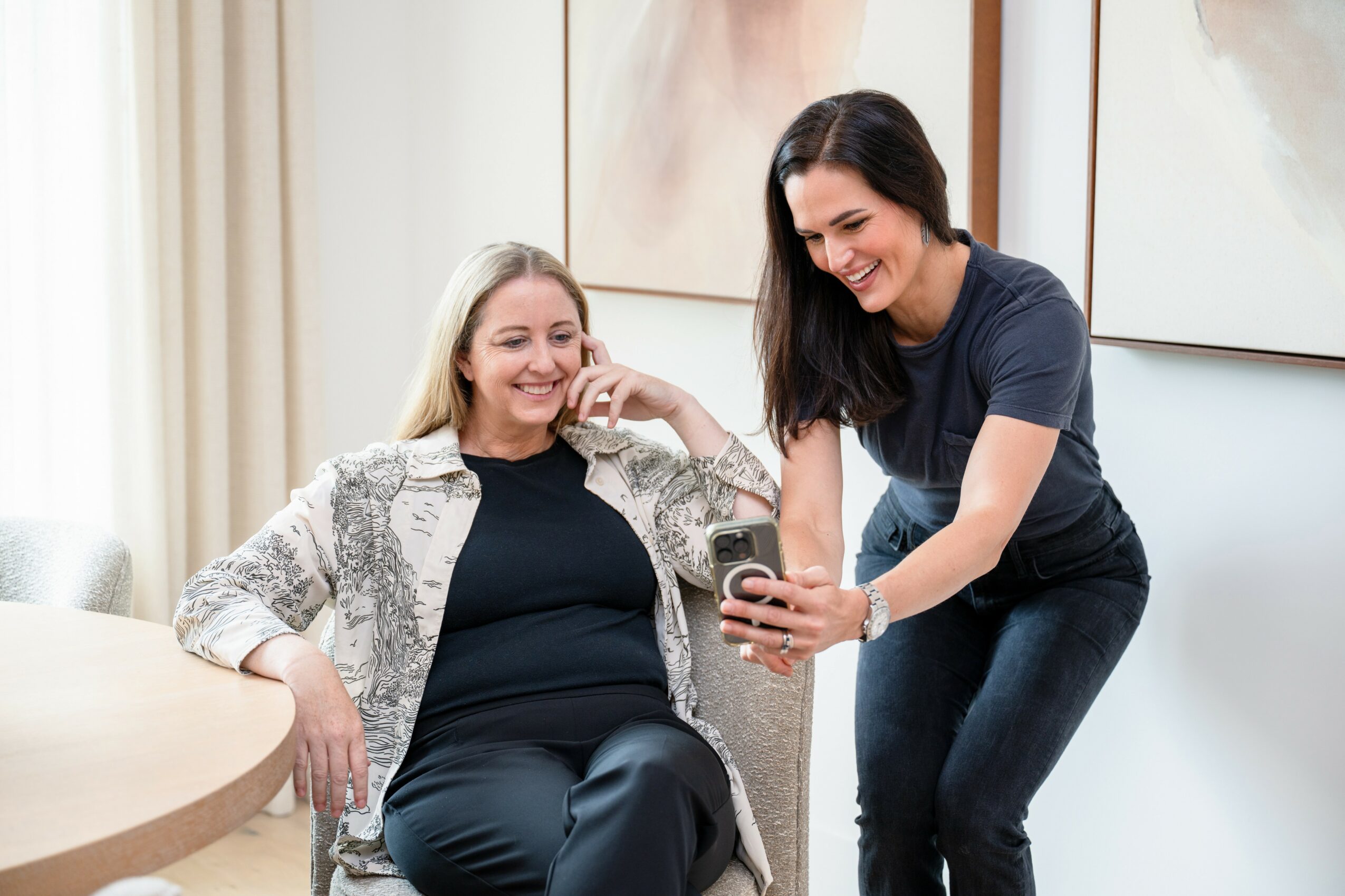Two women smiling and looking at a phone in a bright room with artwork on the wall.