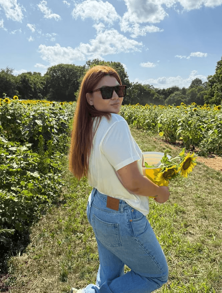 Woman with long hair wearing sunglasses, white shirt, and jeans holding a sunflower in a field under a blue sky.