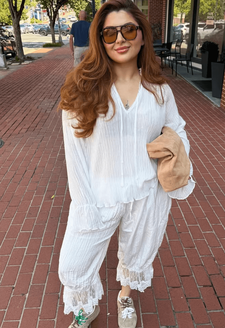 Woman with long wavy hair wearing sunglasses, white outfit, holding a beige bag, standing on brick sidewalk outdoors.