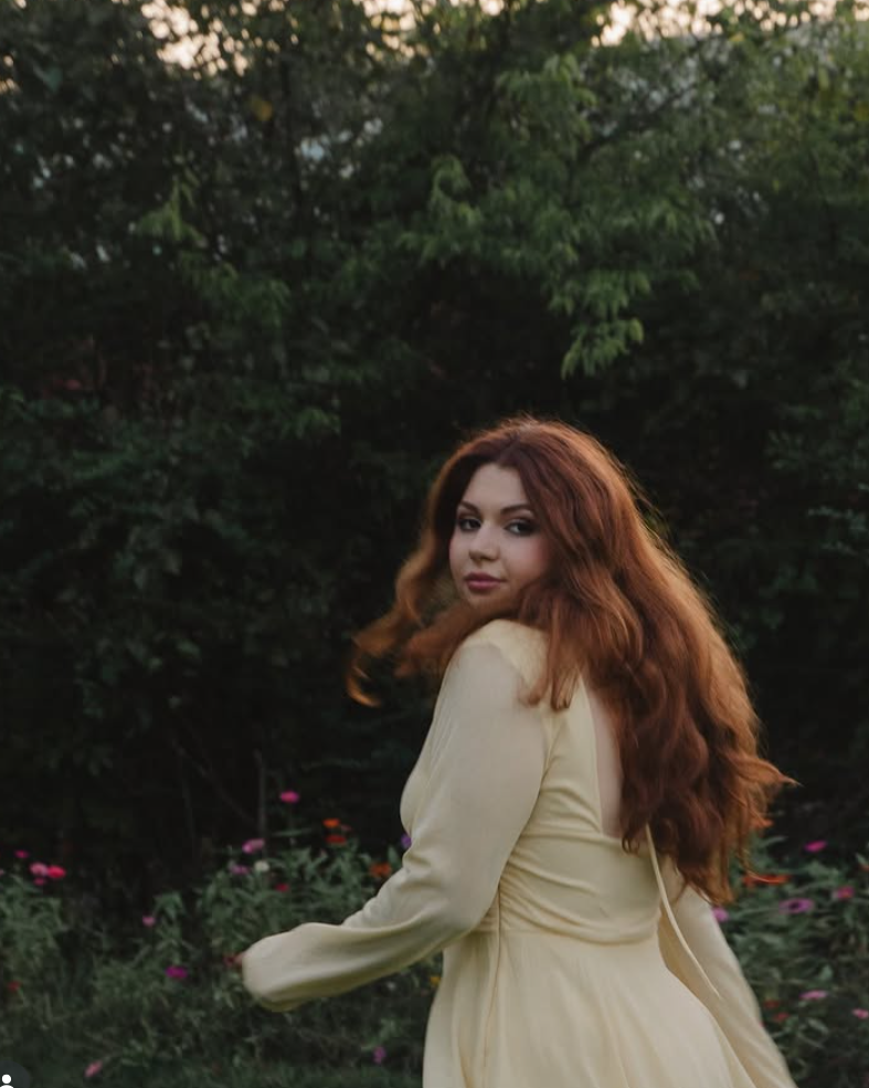 Young woman with long, wavy hair standing outdoors among trees and flowers, looking over her shoulder.
