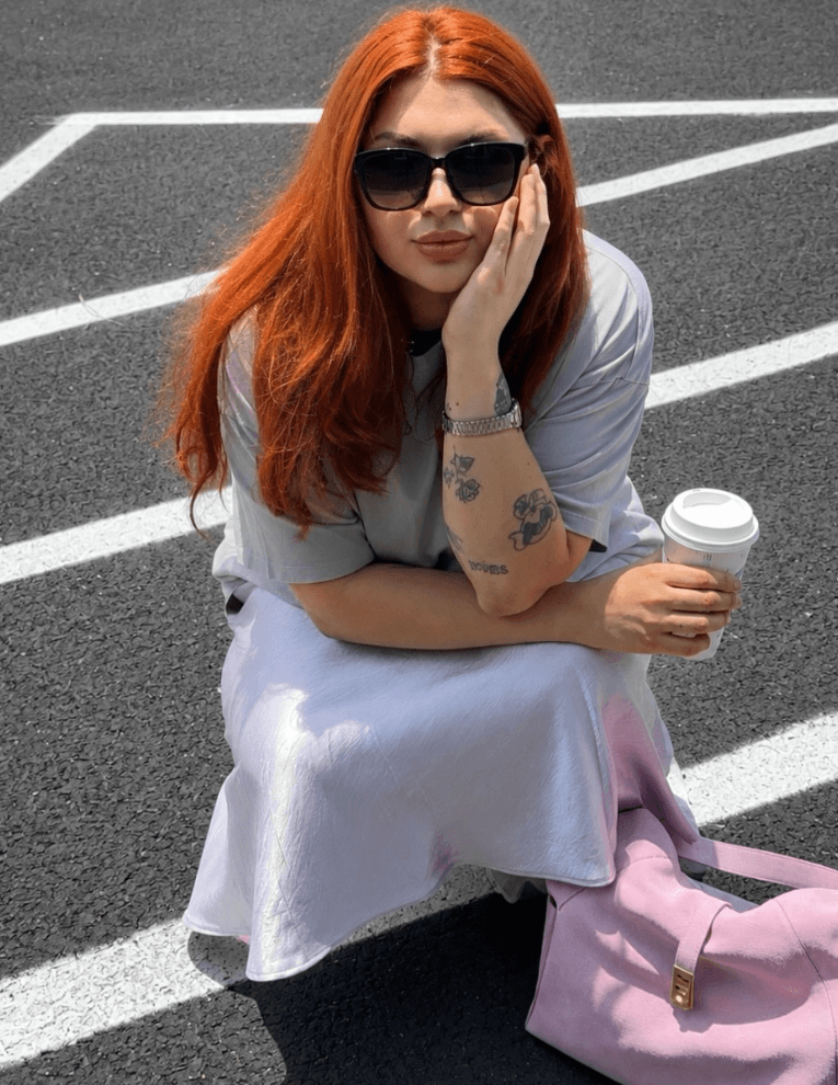 Woman with red hair wearing sunglasses, sitting on the ground with a coffee cup, in a parking lot.