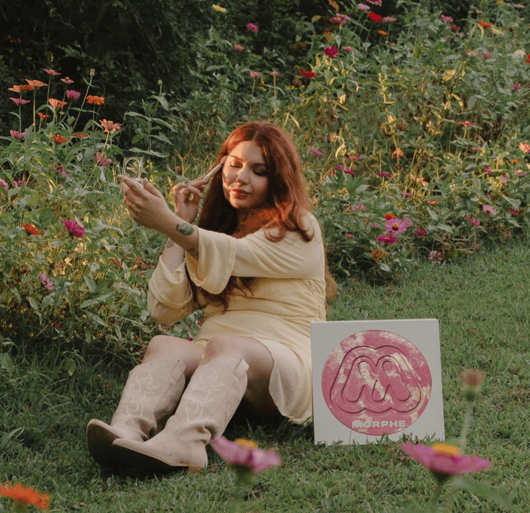 Woman with red hair sitting on grass near flowers, holding a flower, with a pink and white sign nearby.