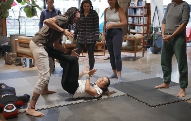 Group of people practicing self-defense techniques with one person on the ground and others assisting in a room.