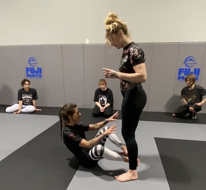 A woman sitting on the mat interacts with a standing woman, with three children sitting against the wall in the background.