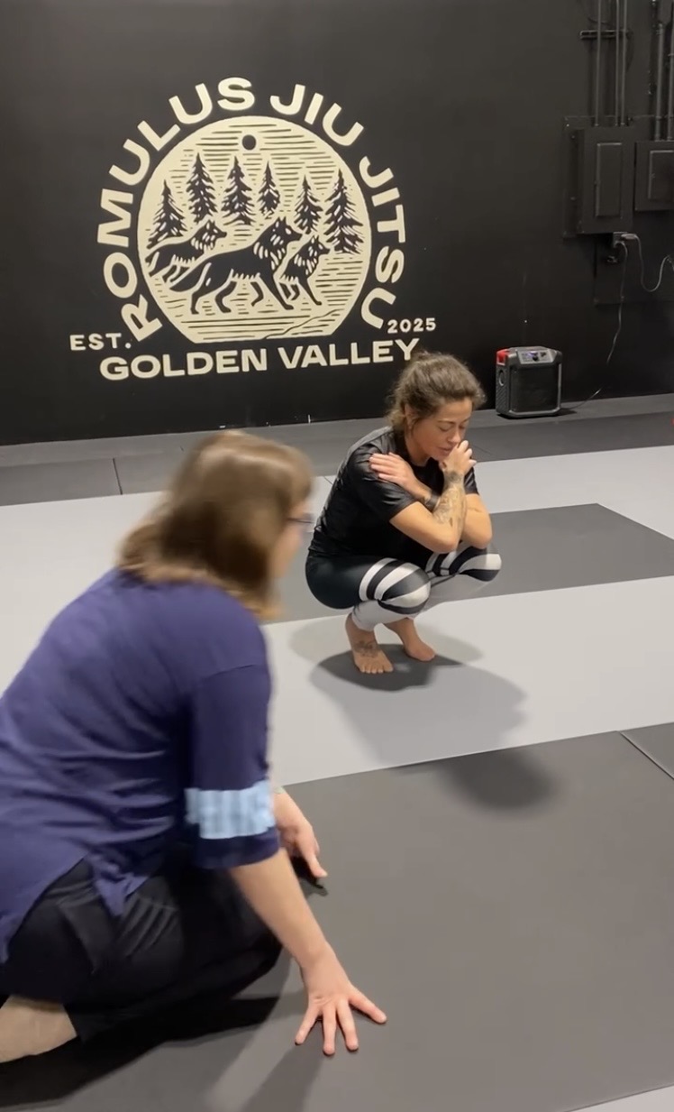 Two children practicing Jiu-Jitsu on mats in a training facility with a logo on the wall behind them, one squatting and the other kneeling.