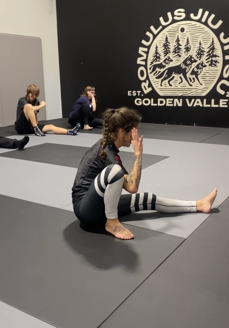 Three people practicing yoga or stretching on mats in a gym with a large circular logo on the wall featuring a wolf and trees.