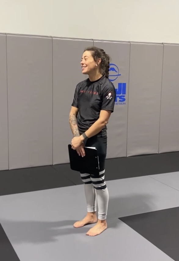 Person standing on a mat, smiling, wearing a black sports shirt and striped leggings, in a gym or training area.