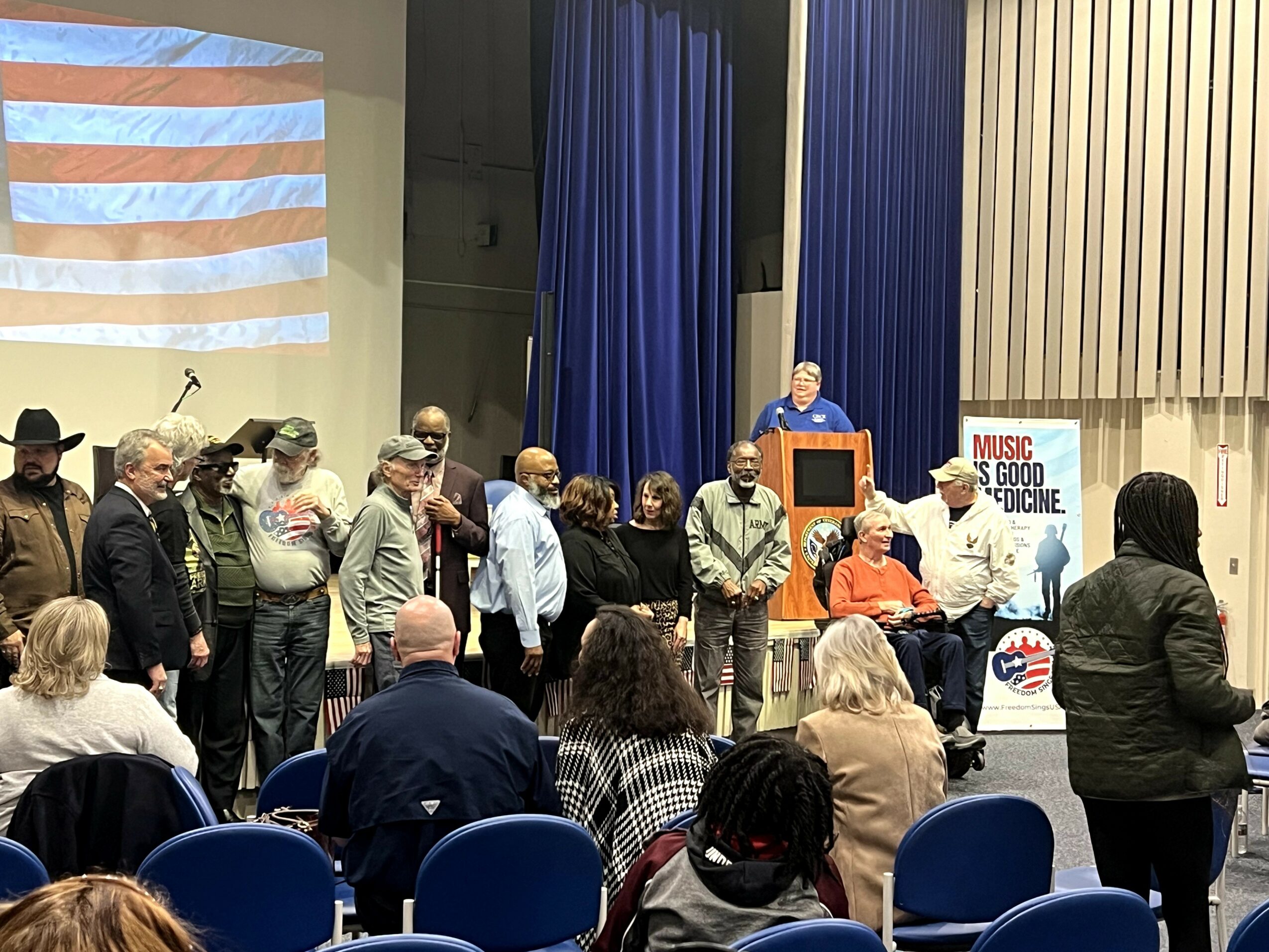 Group of people on stage, some speaking and others listening, with a large American flag projection in the background.