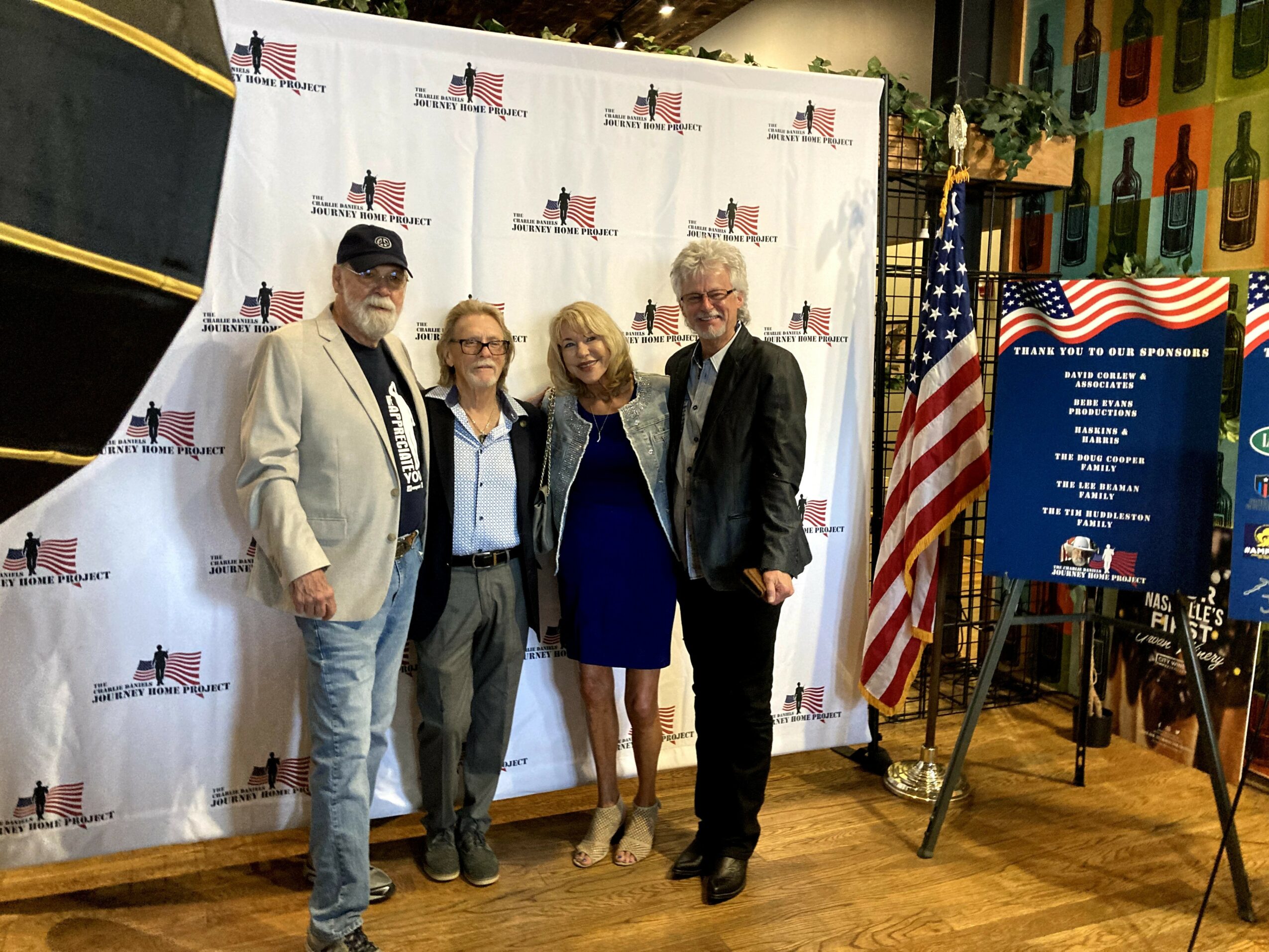 Four people standing in front of a backdrop with American flags and a sign, with an American flag and a blue board on the right.