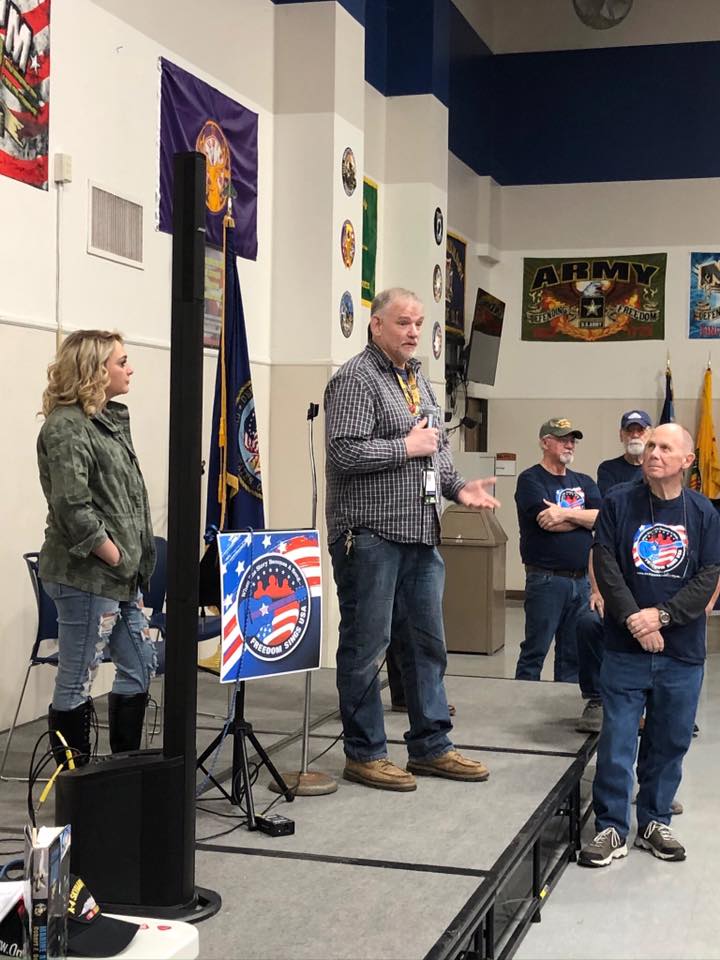 Man speaking into microphone on stage with group of people, flags, and military-themed posters in background.