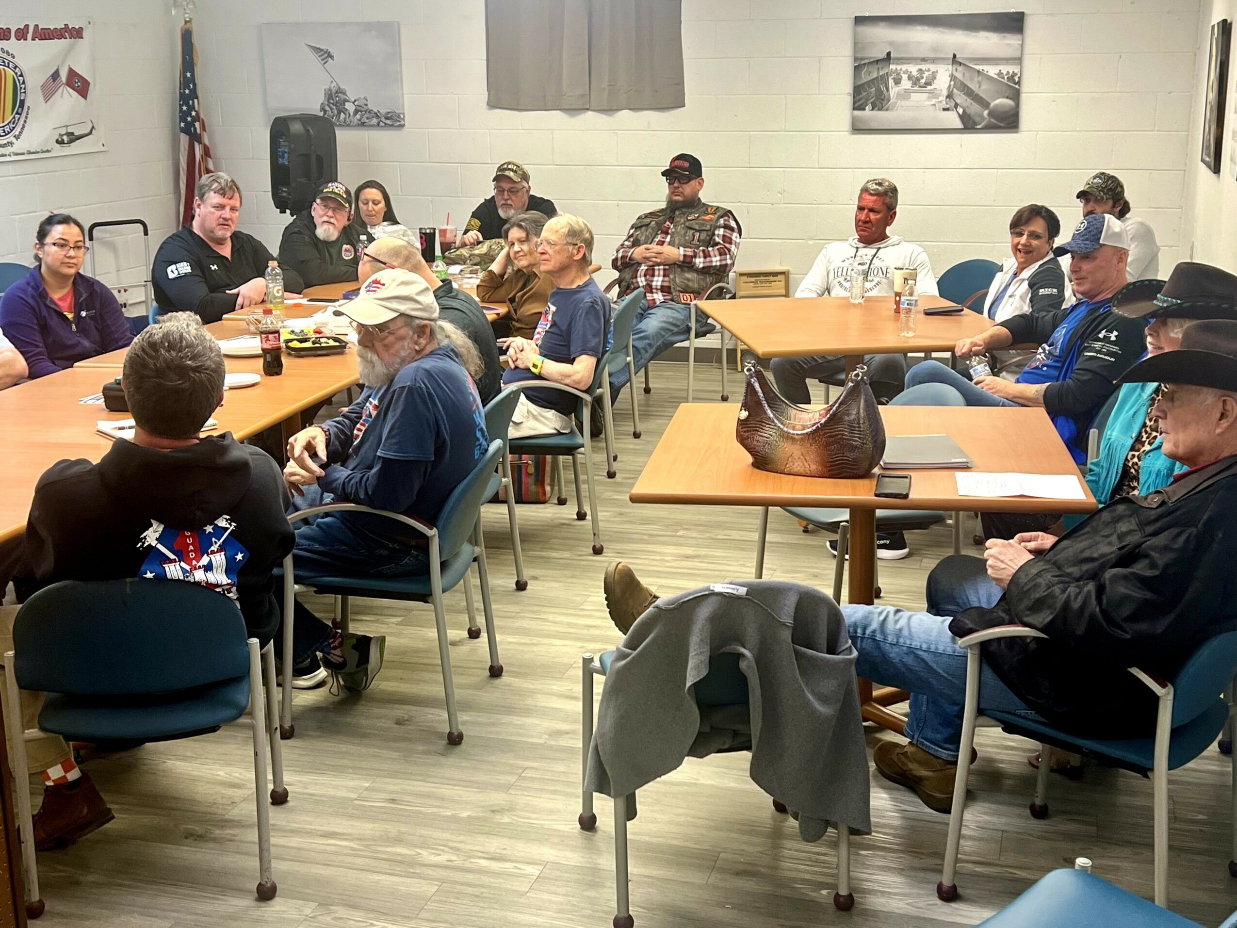 Group of people sitting around tables in a room, engaged in a discussion or meeting, with photographs on the wall.