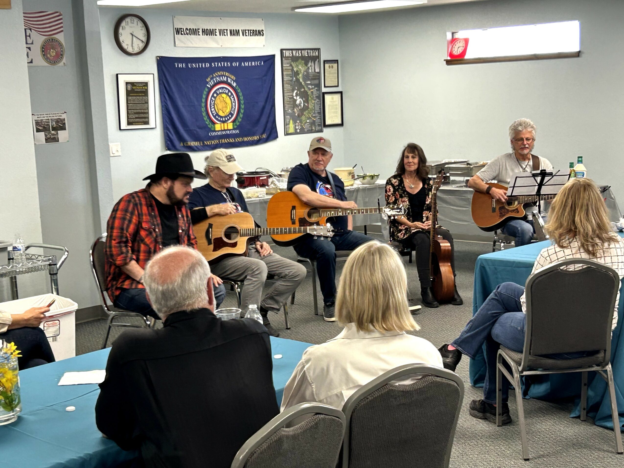 Group of five musicians playing guitars and singing in a room with audience seated at tables, a flag and clock on the wall.