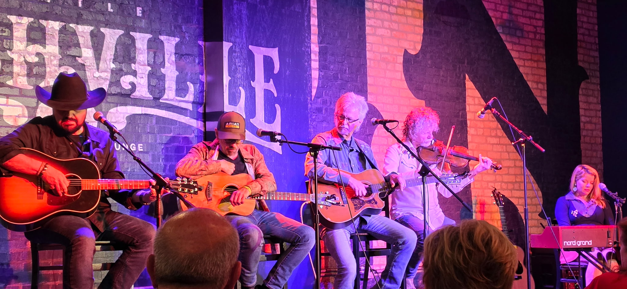 Group of six musicians playing guitars and singing on stage with a colorful background.