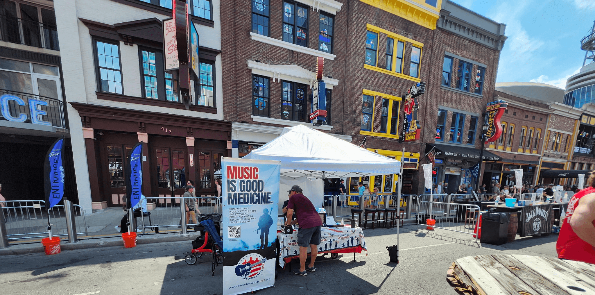 Street scene with a tent selling music merchandise, people browsing, and buildings in the background.