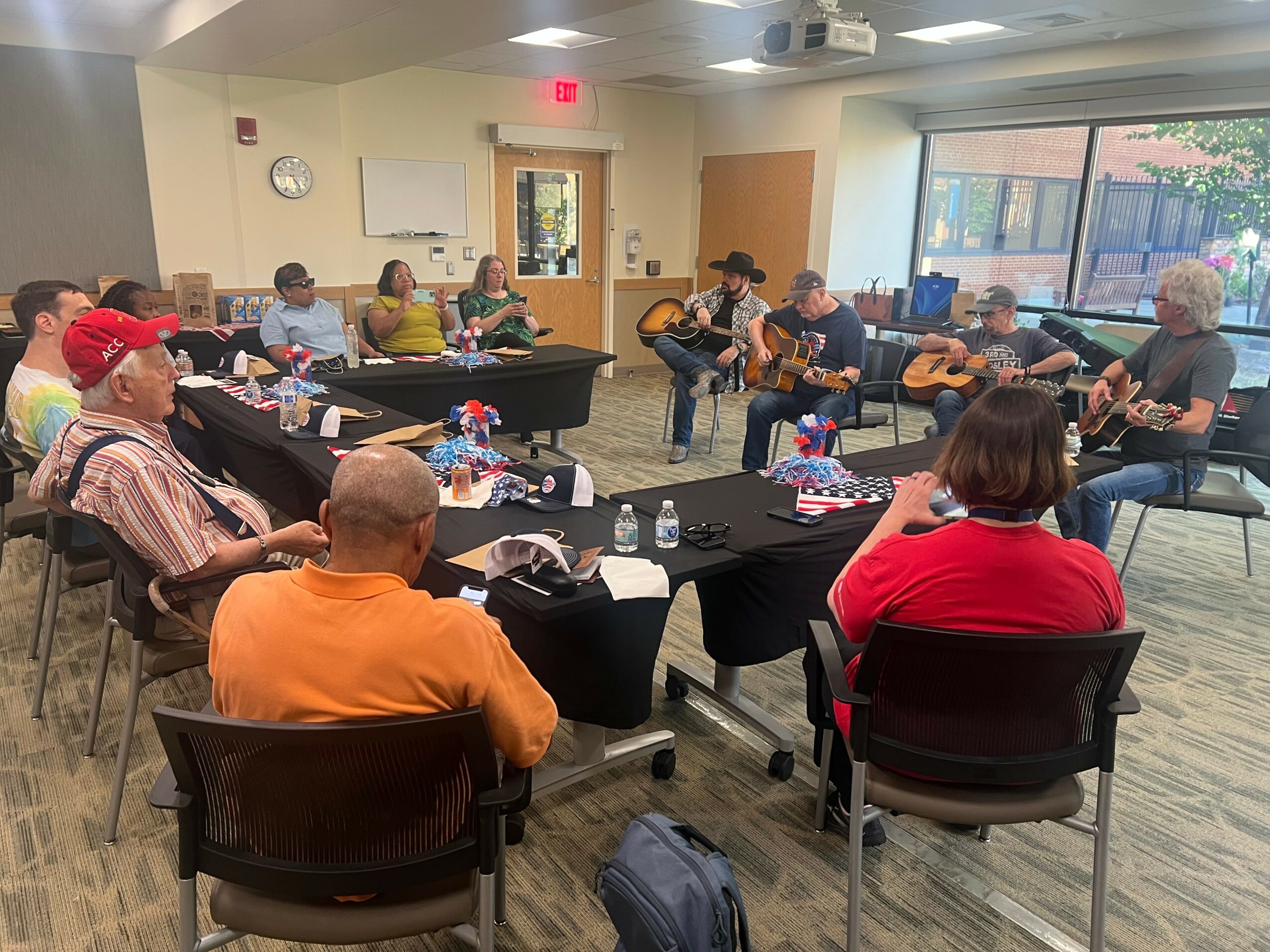 Group of people sitting around tables, some playing guitars, in a room with large windows and a whiteboard.