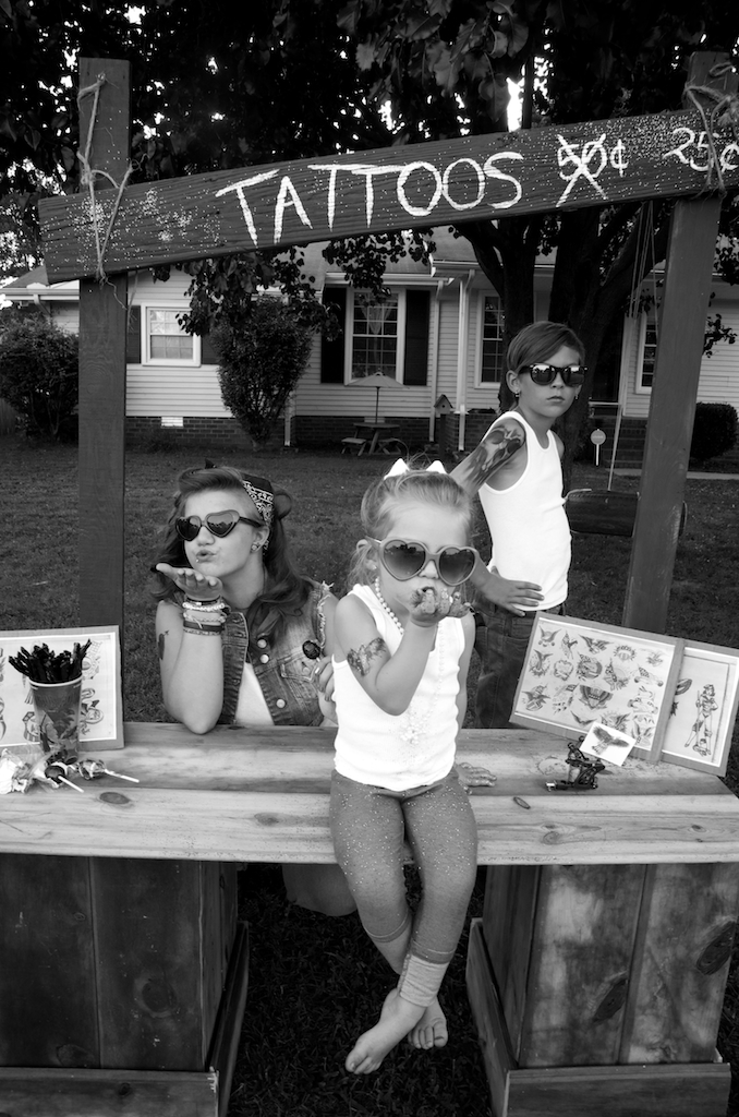 Three children wearing sunglasses at a tattoo stand, with a sign and framed artwork on the table.