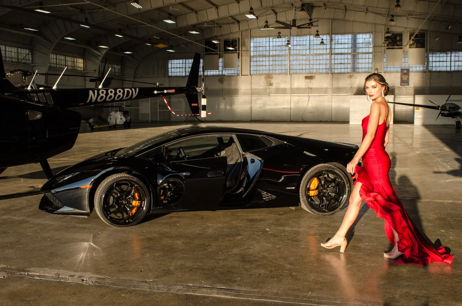 Woman in red gown standing next to a black sports car inside a hangar with a helicopter in the background.