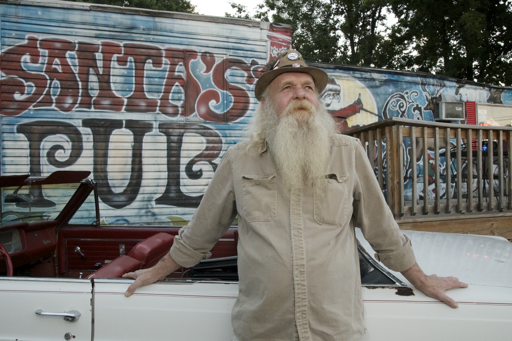 Older man with a long white beard and hat stands outdoors near a graffiti-covered wall, leaning on a vehicle.