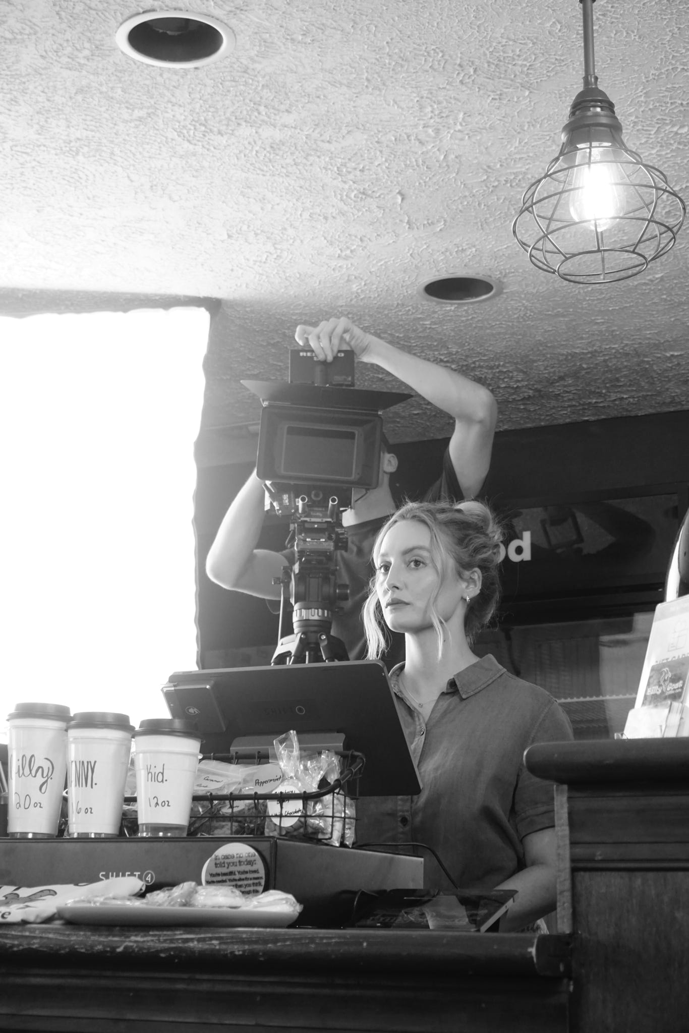 Woman operating camera equipment in a studio with coffee cups on the counter, hanging light, and window in background.