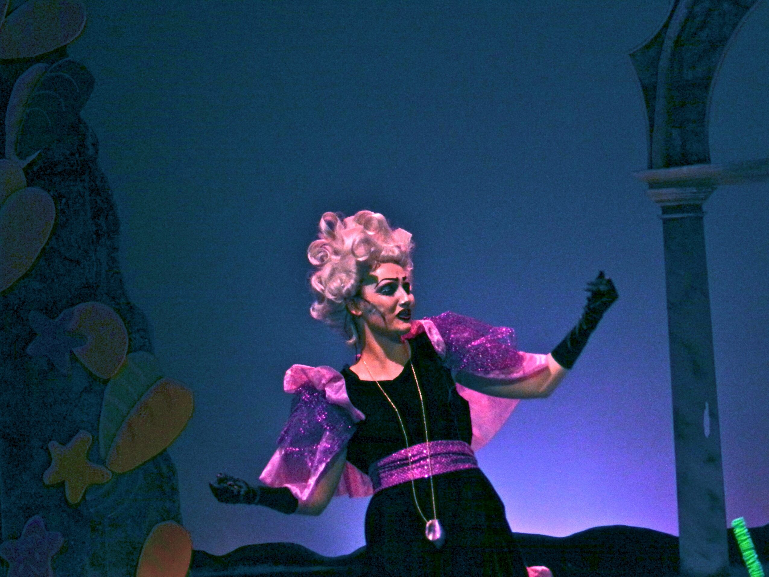 Performer with pink curly hair, wearing purple and black costume, on stage with blue lighting.