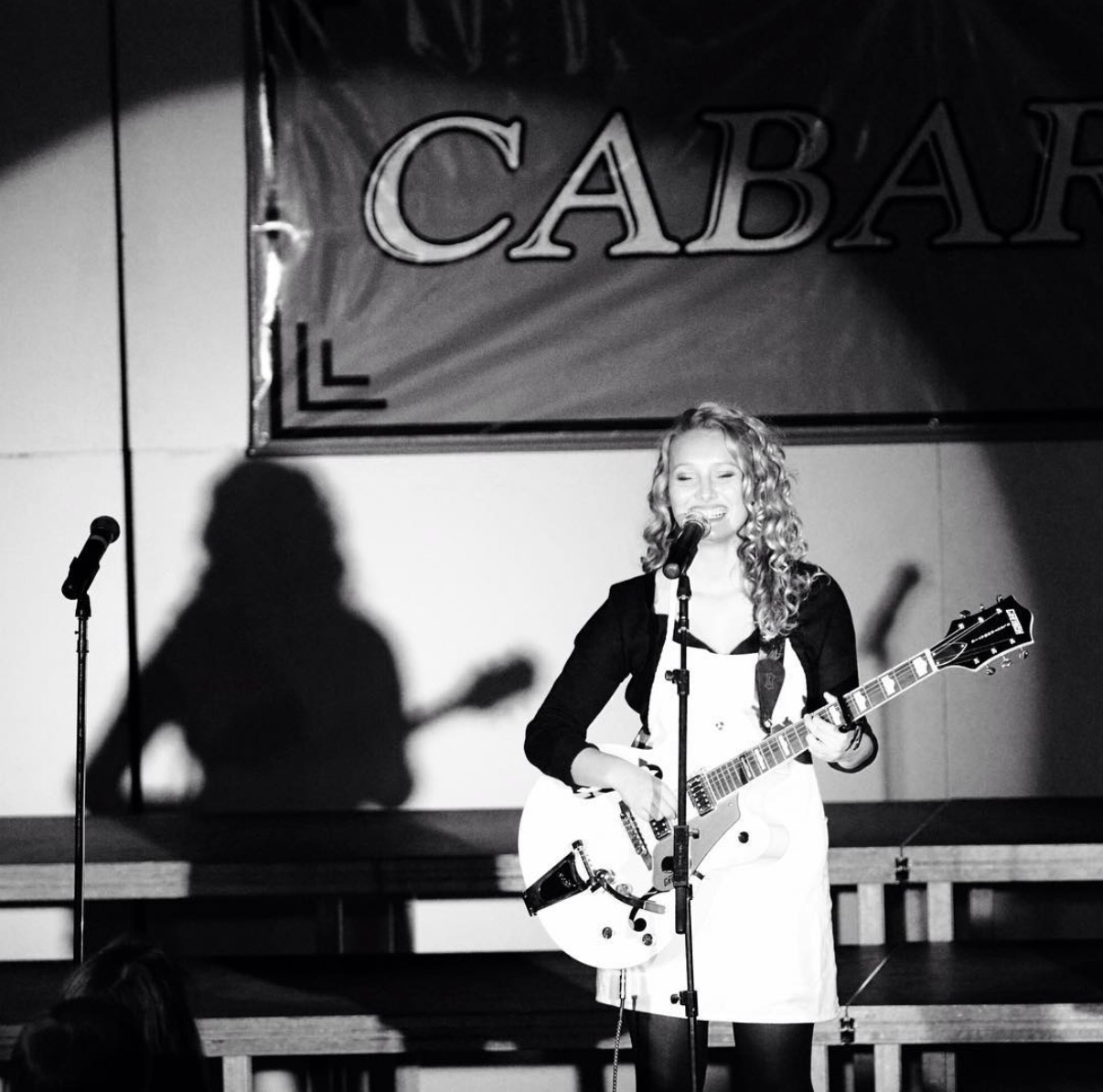 Young woman with curly hair playing guitar and singing on stage, shadow cast on wall, banner with partially visible text behind her.