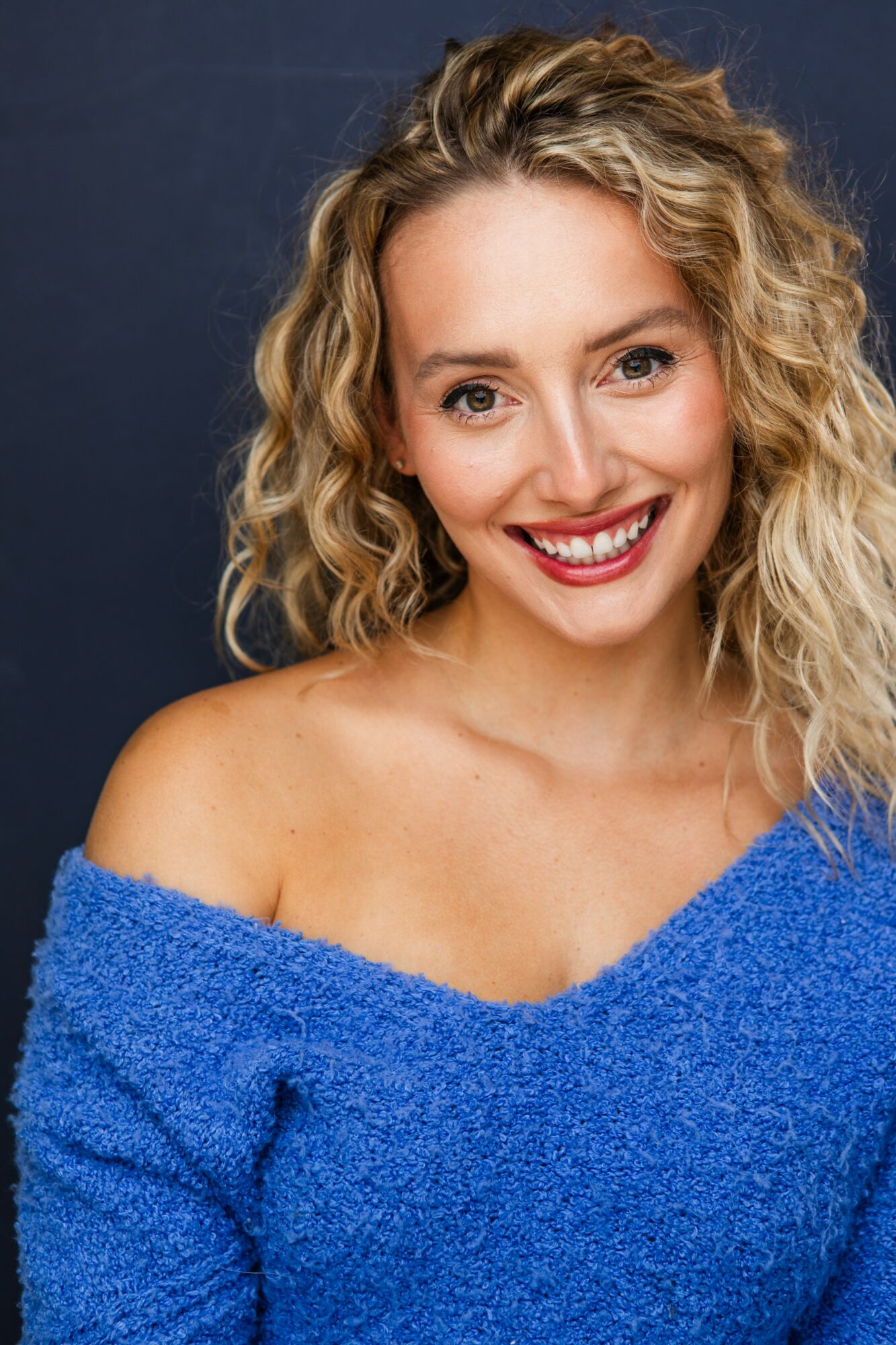 Young woman with curly blonde hair smiling, wearing a blue off-shoulder sweater, against a dark background.