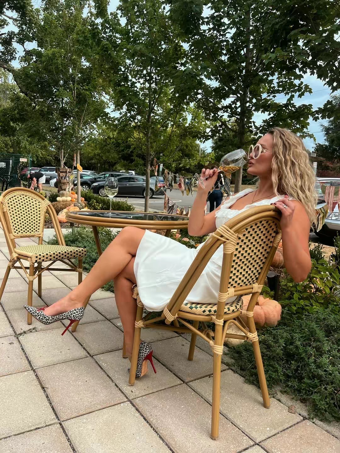 Woman in a white dress sitting outdoors, drinking from a glass, with trees and parked cars in the background.