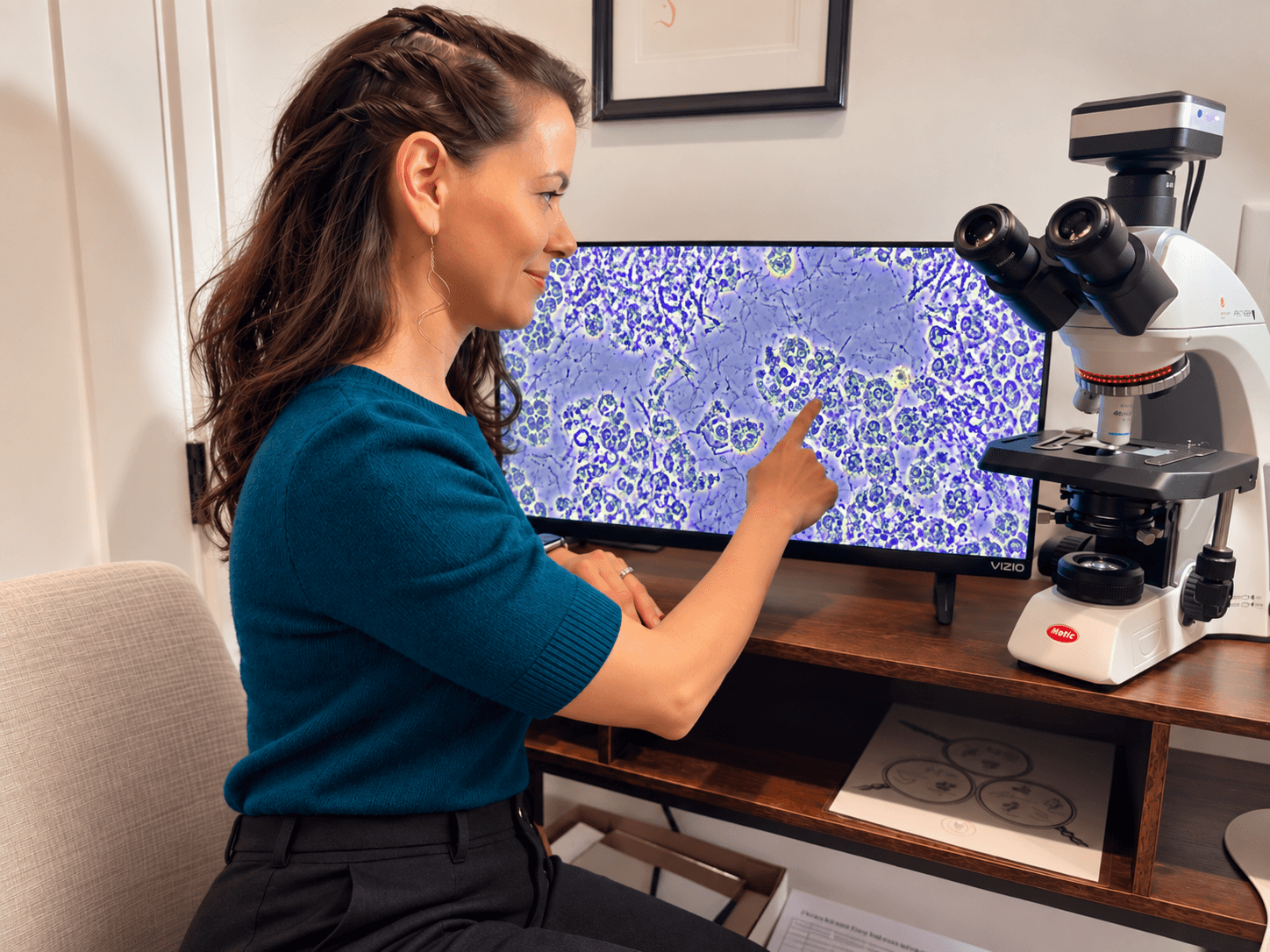 Woman in teal top pointing at a computer monitor displaying microscopic images, with a microscope nearby.