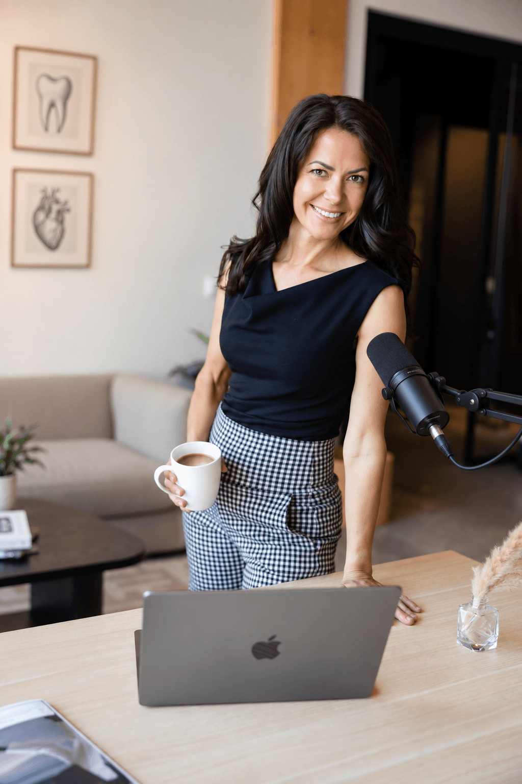 Woman with dark hair smiling, holding a coffee mug, standing at a desk with a laptop, in a modern room.