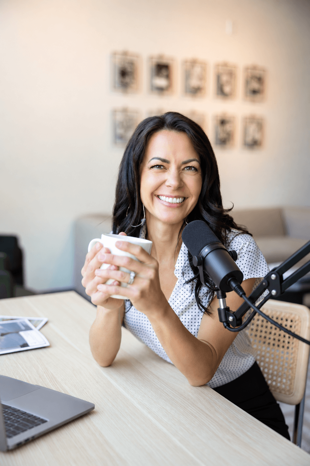 Woman smiling, holding a cup, sitting at a desk with a microphone, in a room with framed pictures on the wall