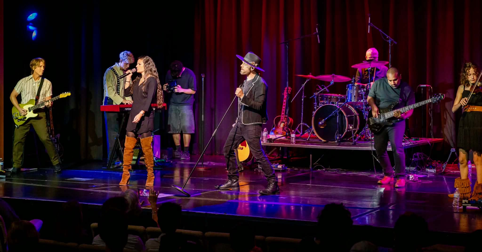 Band performs on stage with red and blue curtains, playing instruments and singing, audience visible in foreground.