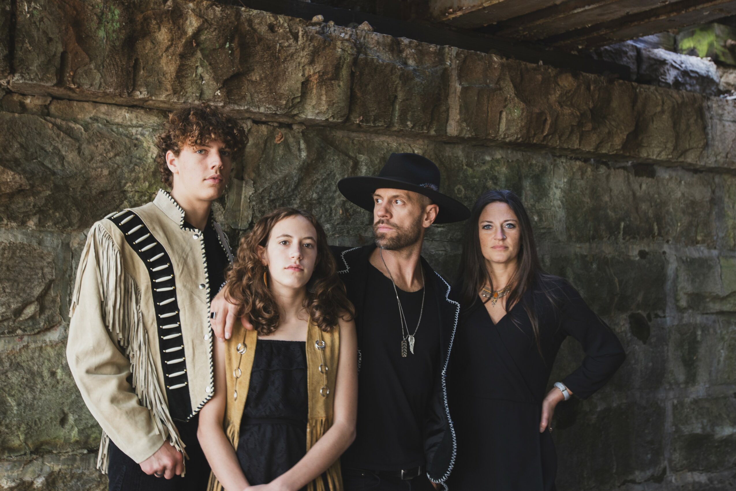 Three people standing against a stone wall, one wearing a hat, all looking in different directions.
