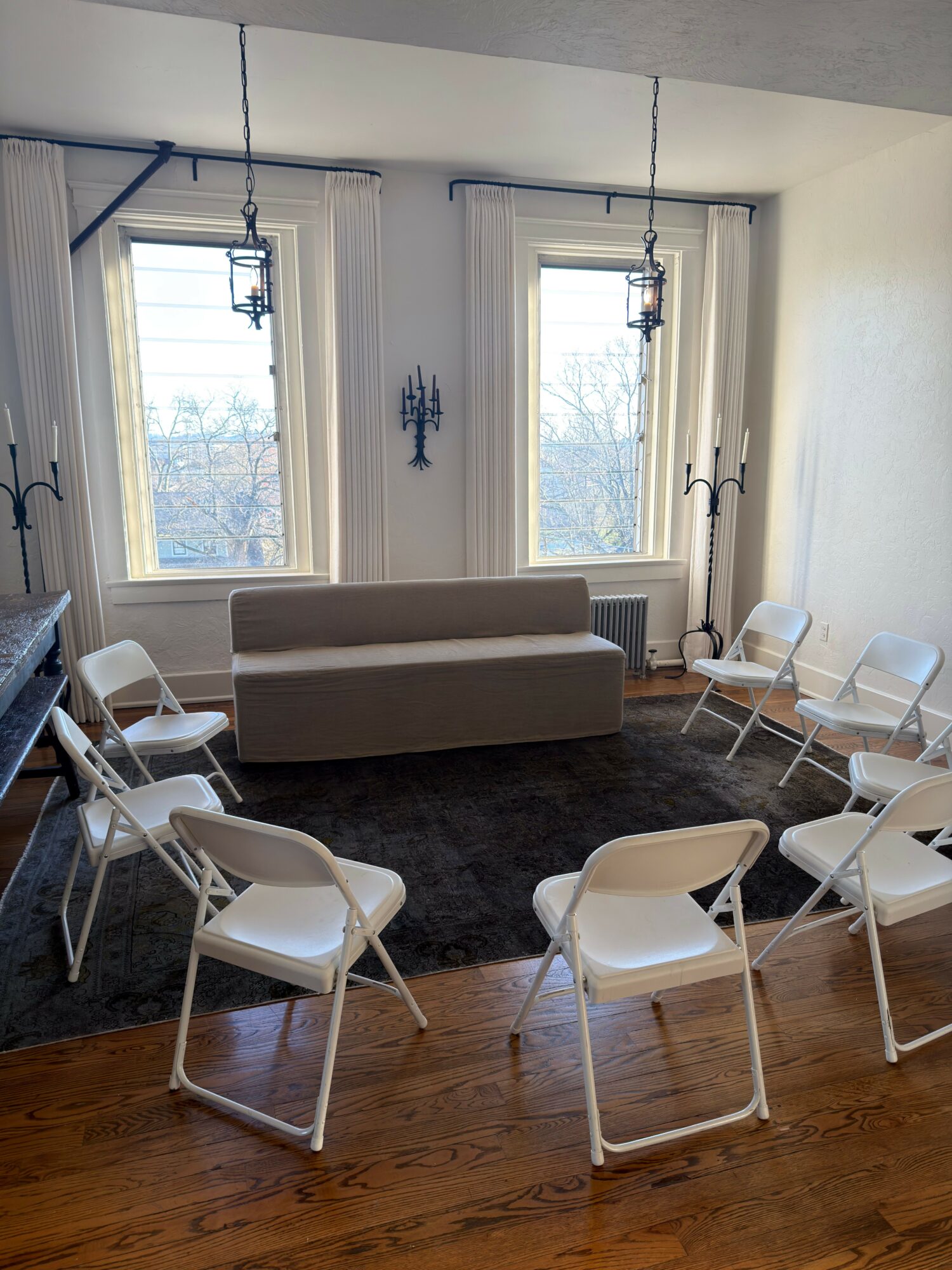 Room with two large windows, a sofa, and several white chairs on a wooden floor.
