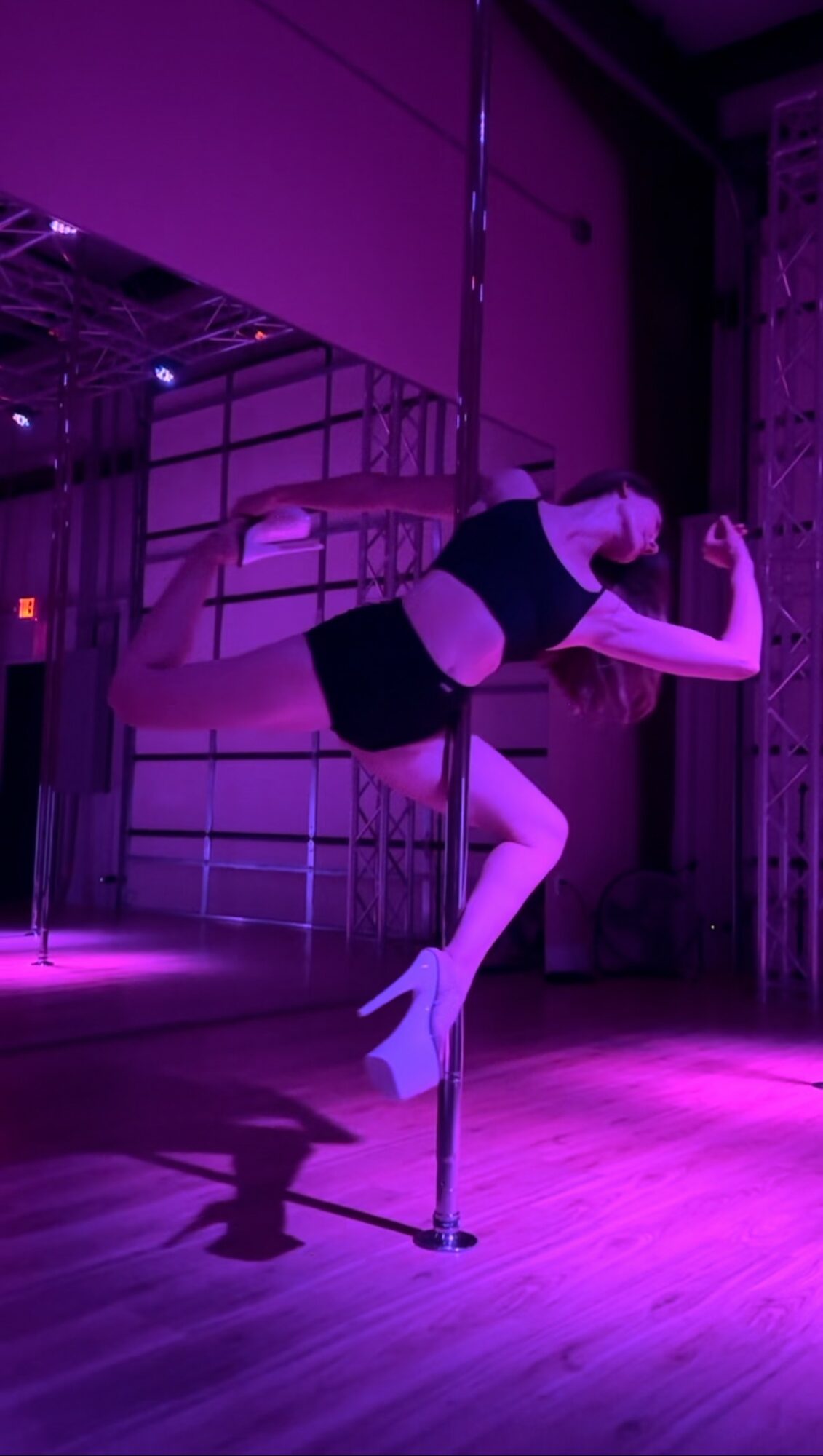 Performer in black outfit poses on pole in purple-lit stage, one leg extended, arms flexed, with stage equipment in background.
