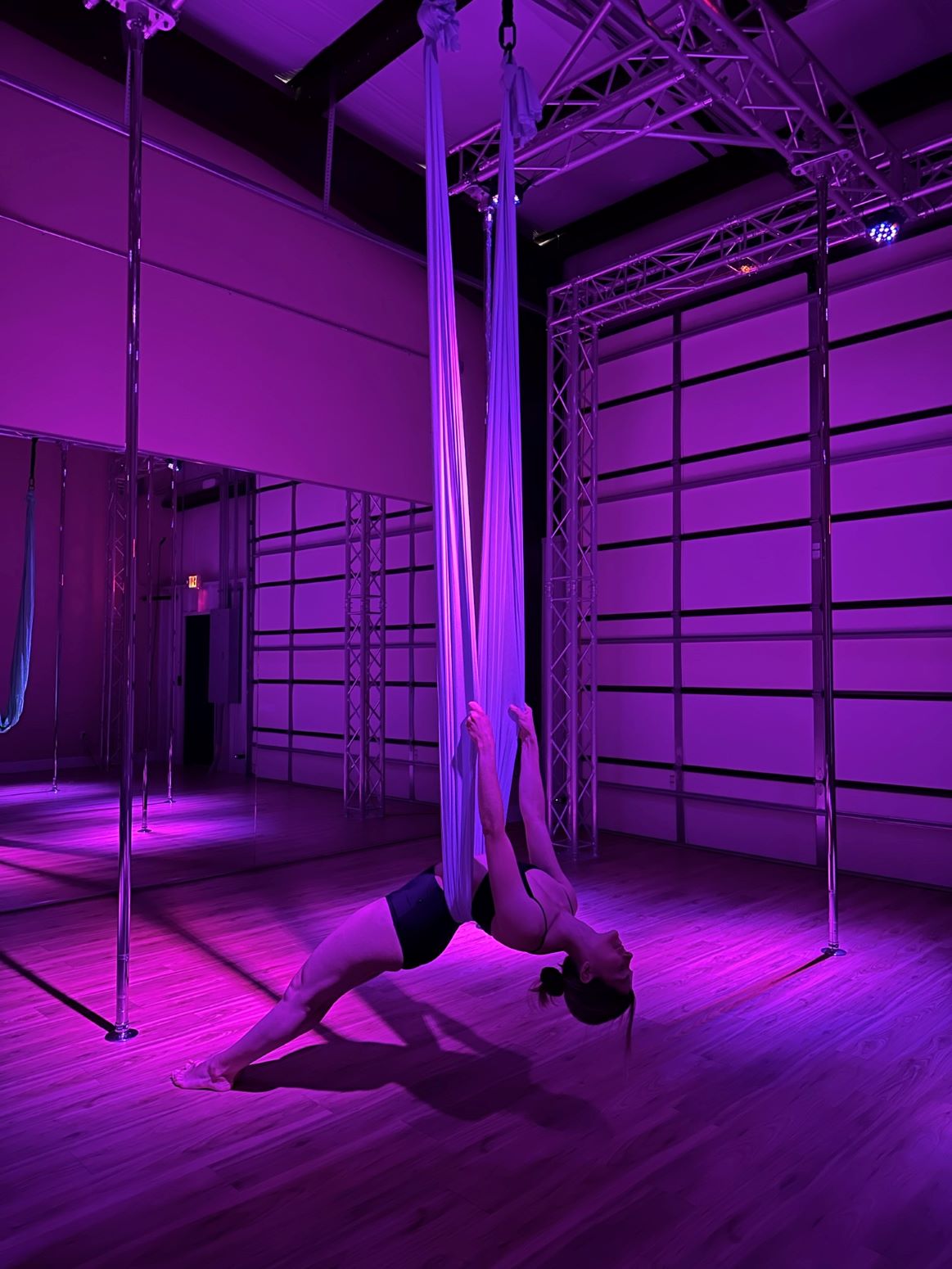 Person practicing aerial silk dance in purple-lit studio, holding fabric with legs extended upward, on wooden floor.