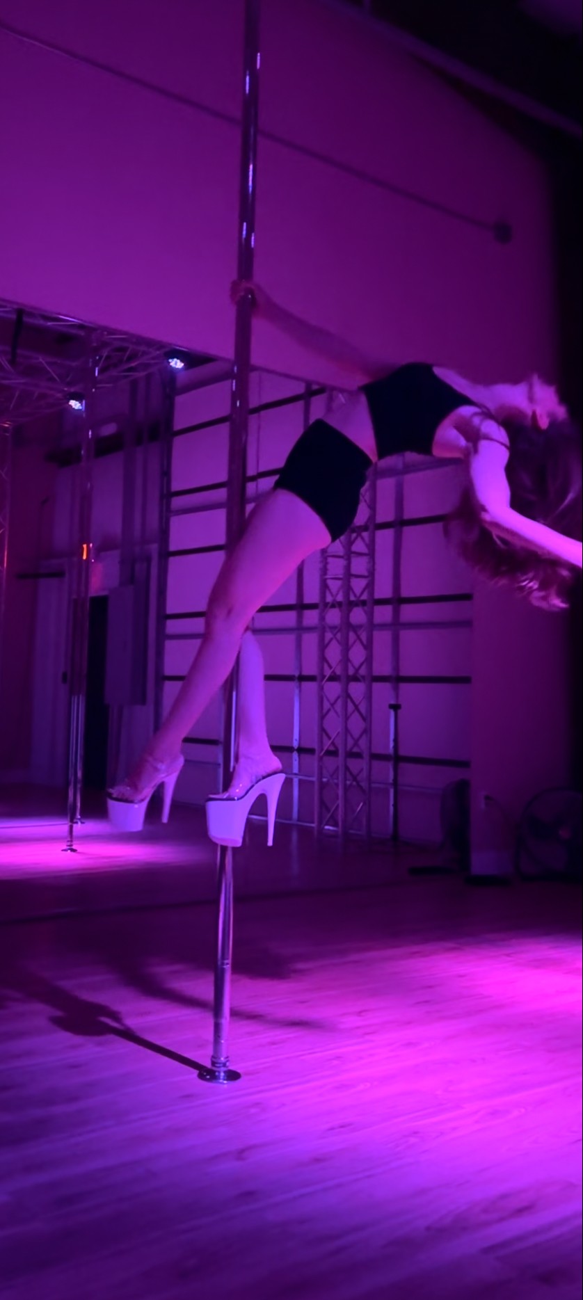 A woman performs pole dance in a dimly lit studio with purple lighting, wearing black shorts and high heels.