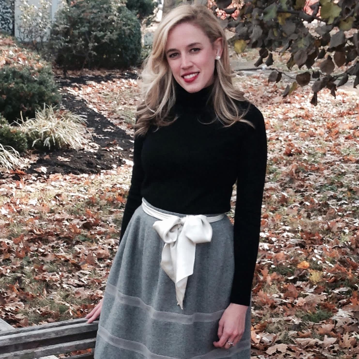 Young woman with blonde hair in a black turtleneck and gray skirt outdoors among fallen leaves.