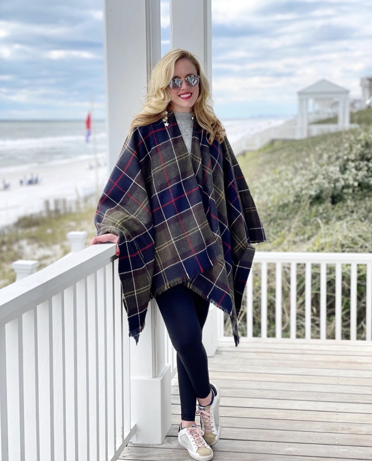 Woman with blonde hair and sunglasses standing on a porch with a beach view, wearing a plaid poncho, dark pants, and sneakers.