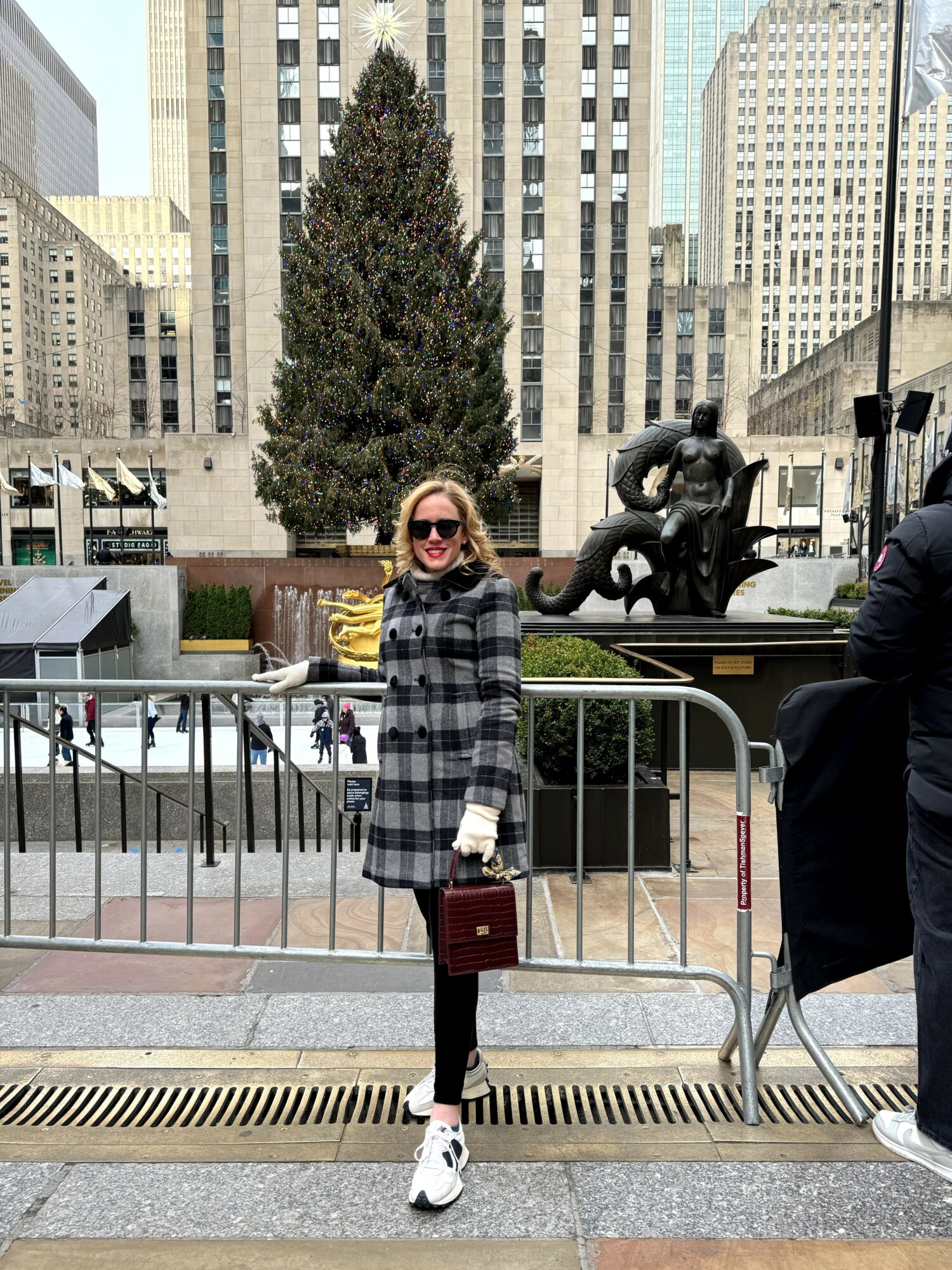 Woman with sunglasses and a checkered coat standing outdoors in an urban area with tall buildings and a statue behind her.