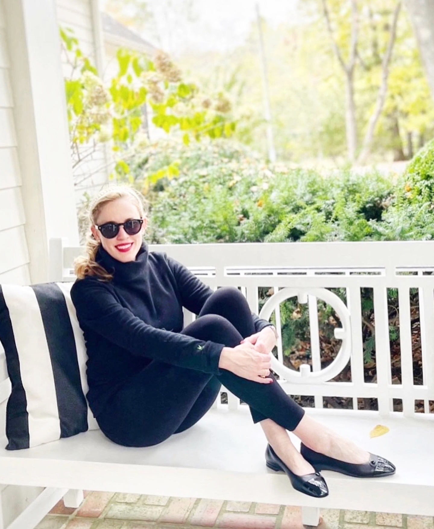 Woman with sunglasses sitting on a white bench outdoors, surrounded by greenery, wearing a black outfit and smiling.
