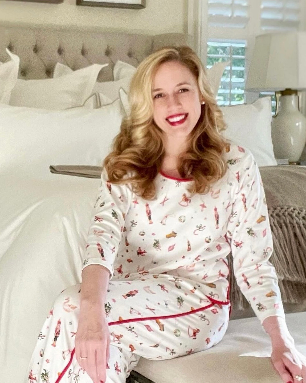 Woman with long wavy hair in pajamas sitting on bed, smiling, in a bedroom with white bedding and lamps.