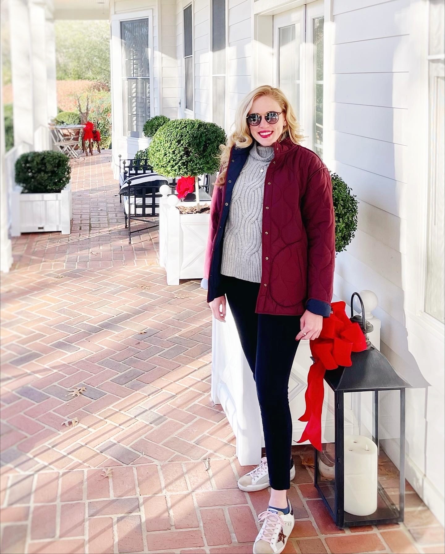 Woman with sunglasses and red jacket standing outside near white building, potted plants, and lantern, smiling.