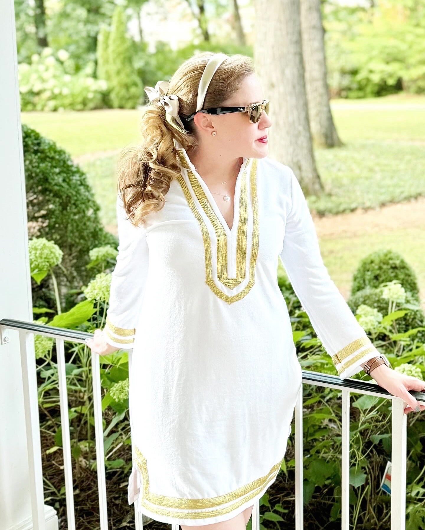 Woman in white dress with gold accents standing outdoors near a railing, surrounded by greenery and trees.