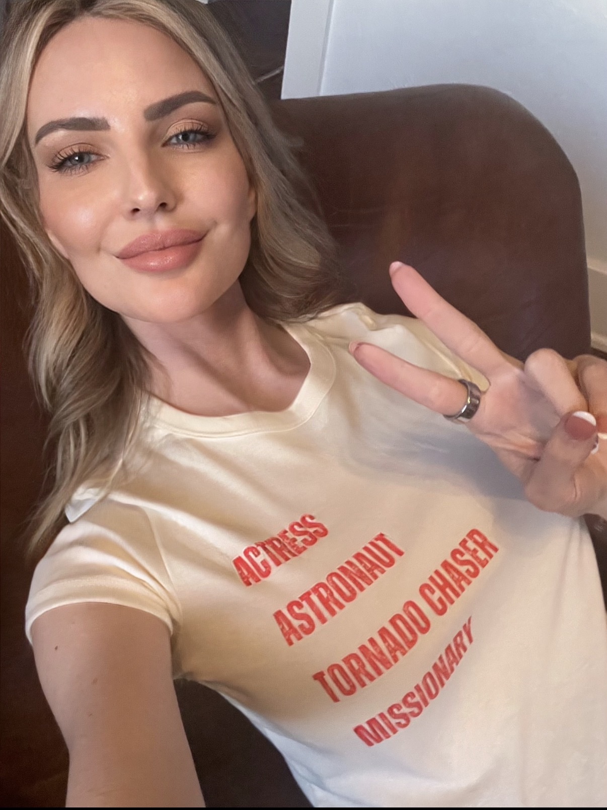 Young woman with blonde hair making a peace sign, wearing a beige T-shirt with red text, smiling.