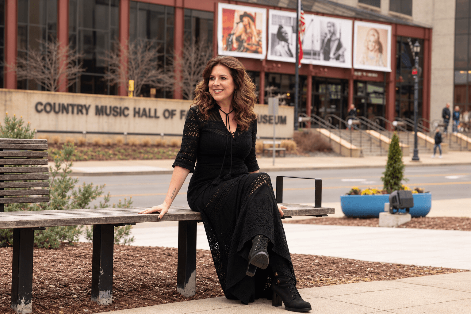 Woman with curly hair sitting on a bench outside, smiling, wearing a black dress and boots, in an urban setting.