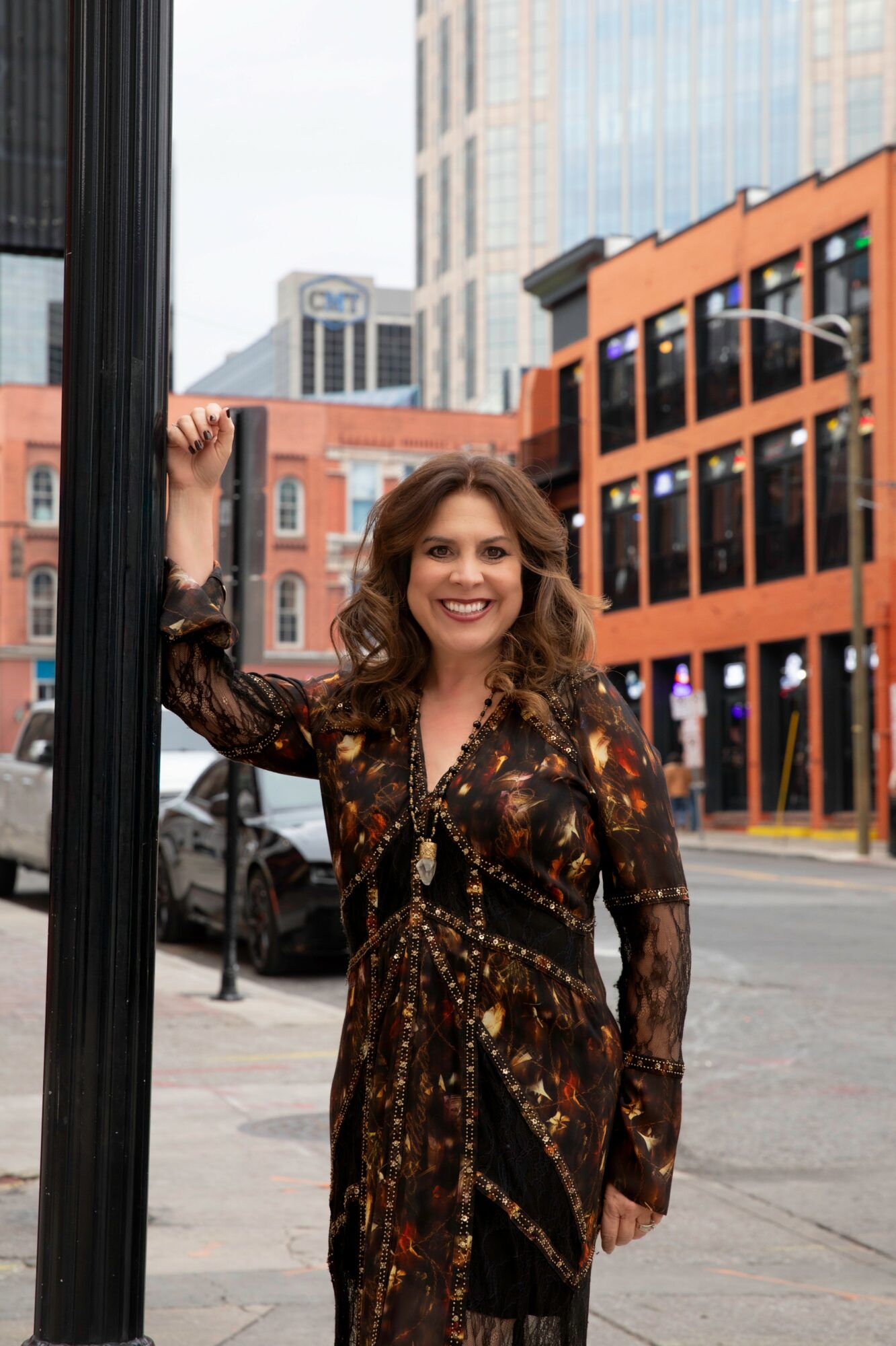 Woman with curly hair smiling, standing outdoors in an urban area with buildings and cars in the background.