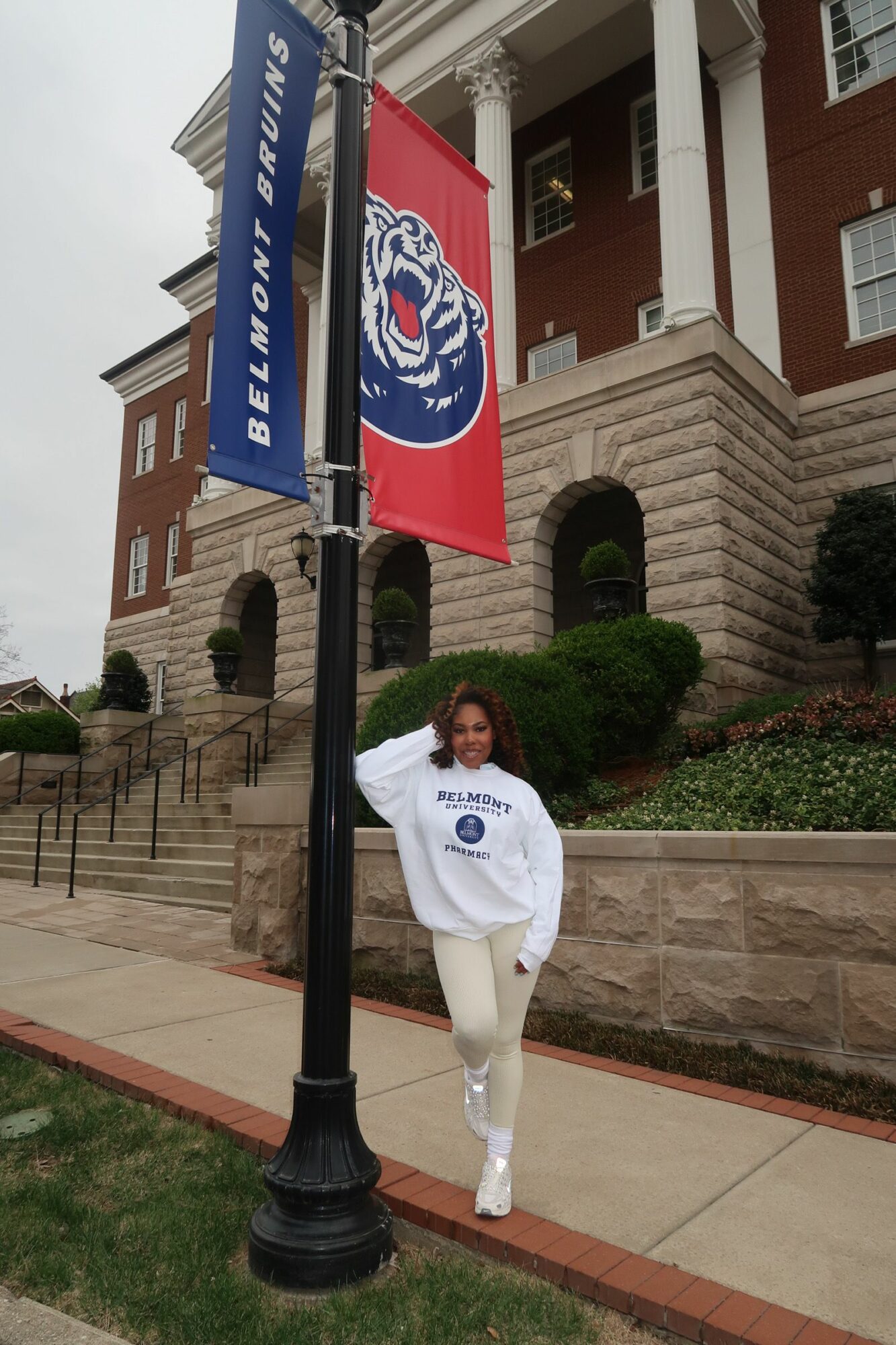 A person in a white martial arts uniform standing on a sidewalk near a flagpole with a blue banner, in front of a building with stairs.
